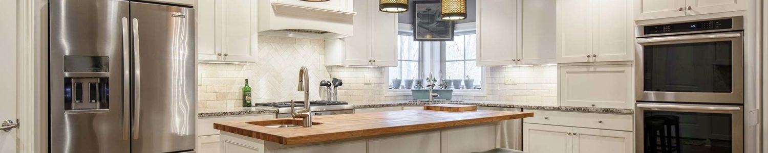 Wide view of the renovated kitchen in the Gillispie Residence, highlighting the spacious wood-topped island, extensive white cabinetry, and modern stainless steel appliances including double ovens.