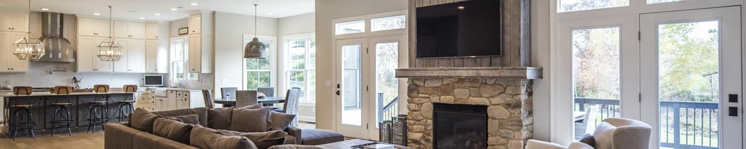 Large great room featuring a stone fireplace with a wooden mantel and integrated media space, with double doors leading to an outdoor deck, connecting to the kitchen in the background.