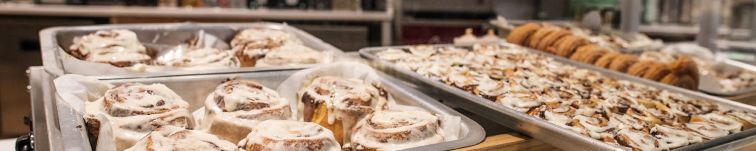 Display counter at Cinnabon showcasing trays of freshly baked goods, with various coffee machines and service equipment in the background.