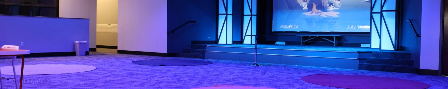 Main event space with a raised stage, large projection screen, and integrated lighting, featuring a patterned carpeted floor.